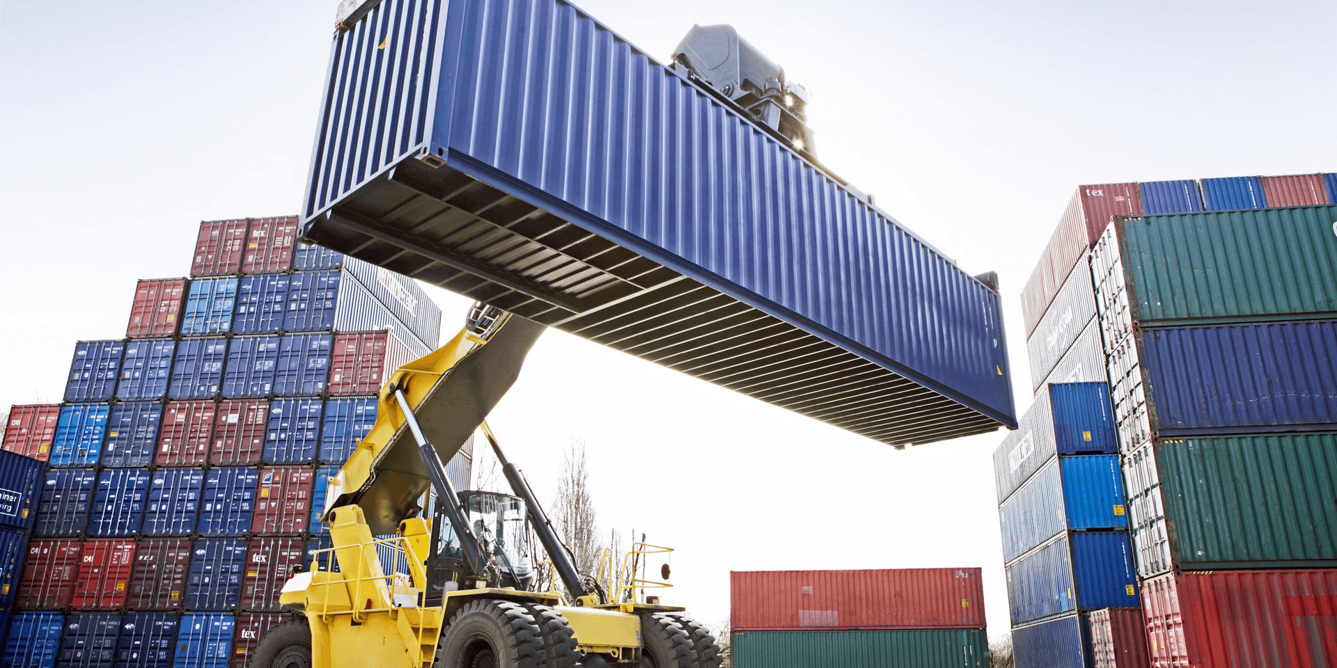 A yellow crane lifting a large blue shipping container among stacked containers at a port.