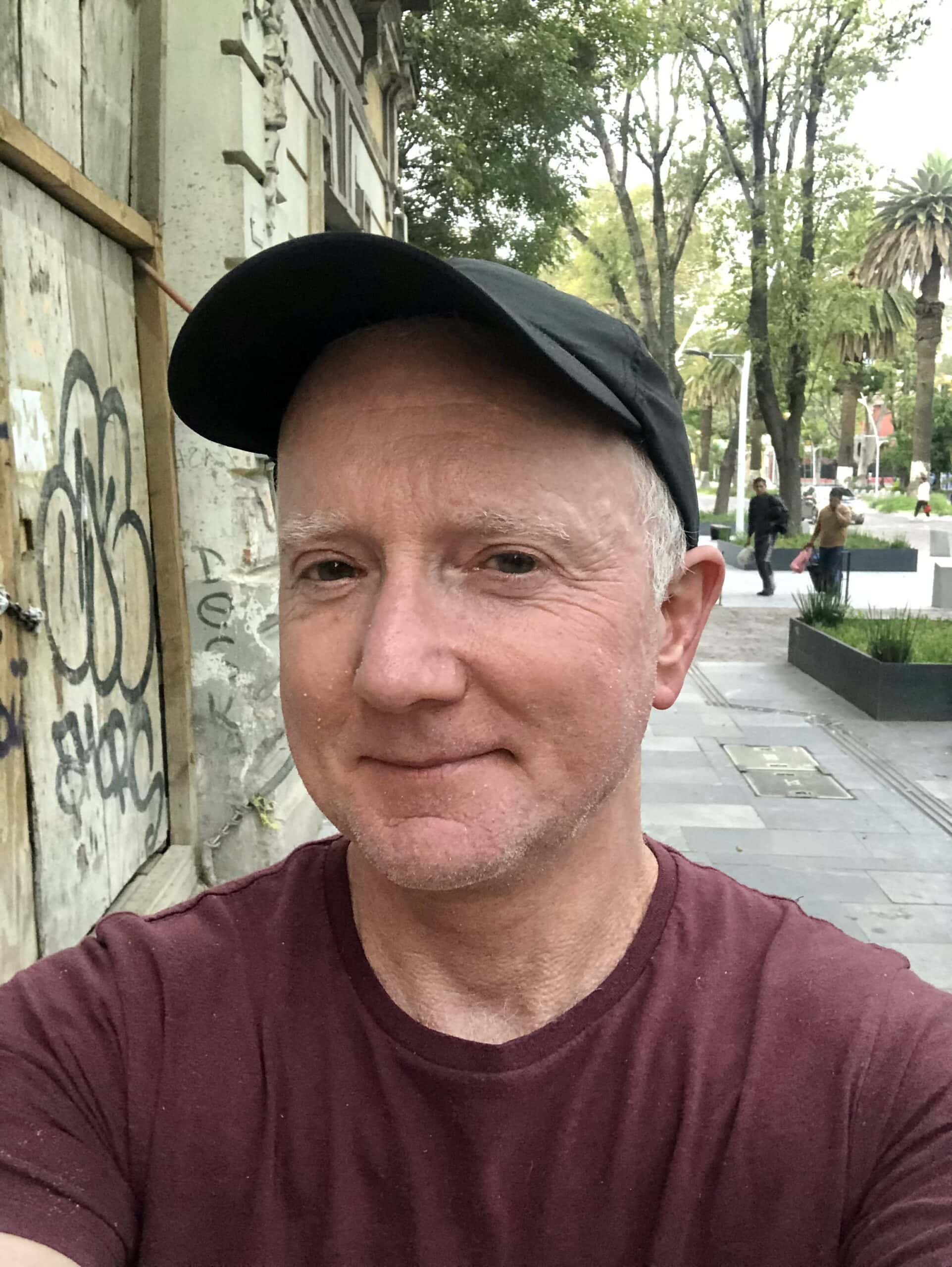 Smiling man taking selfie outdoors, urban street scene with trees, graffiti wall, and pedestrians in the background, casual fashion with cap and maroon t-shirt, relaxed mood.