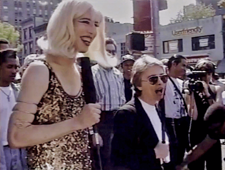 People gathered in an outdoor urban setting during a daytime event with media coverage, including a woman holding a microphone and a man with sunglasses, showcasing lively street scene and crowd engagement.