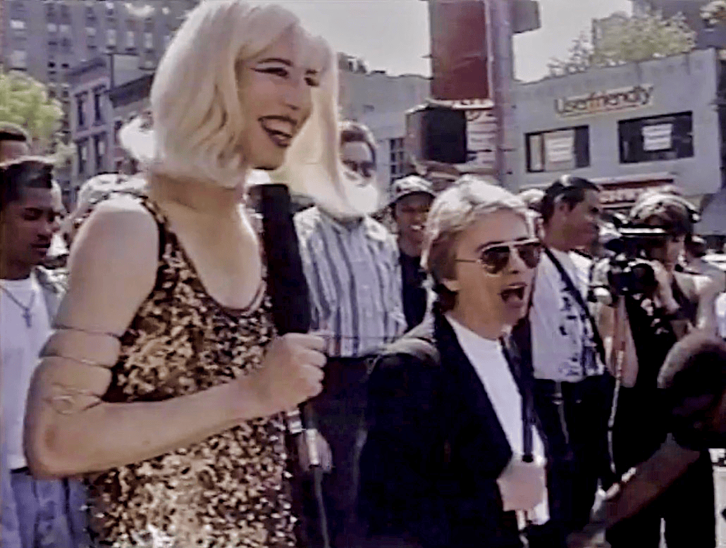 People gathered in an outdoor urban setting during a daytime event with media coverage, including a woman holding a microphone and a man with sunglasses, showcasing lively street scene and crowd engagement.
