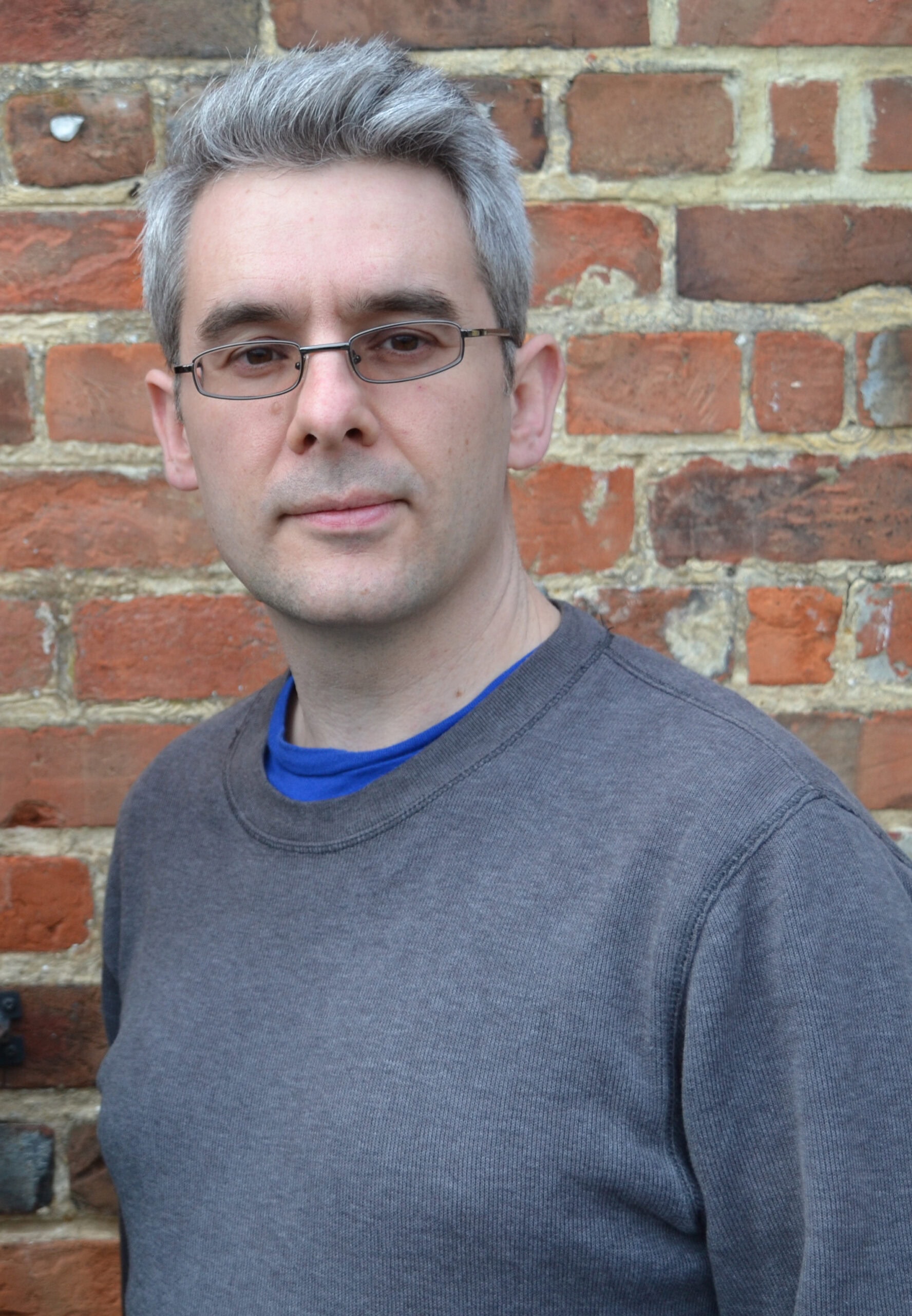 Portrait of a man wearing glasses in front of a brick wall, showcasing a casual and confident appearance suitable for professional or social context.