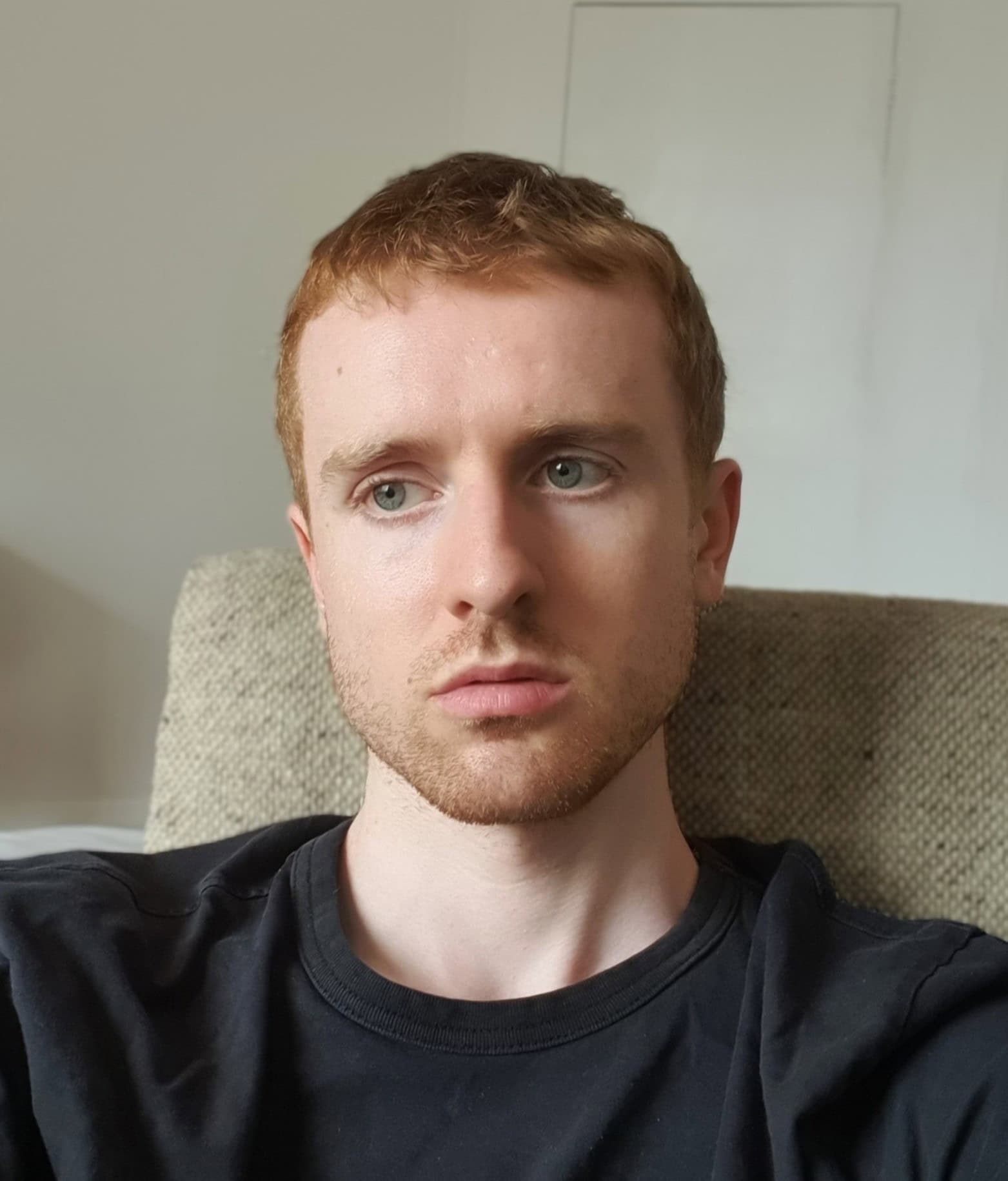 Young man with red hair and blue eyes relaxing on a sofa, casual black T-shirt, natural indoor lighting, modern interior, lifestyle, portrait, comfortable home setting.
