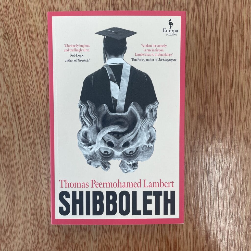 Vintage book cover of "Shibboleth" by Thomas Peermohamed Lambert, featuring an artwork of a person in a graduation cap and gown with a stylised dragon illustration, on a wooden surface.