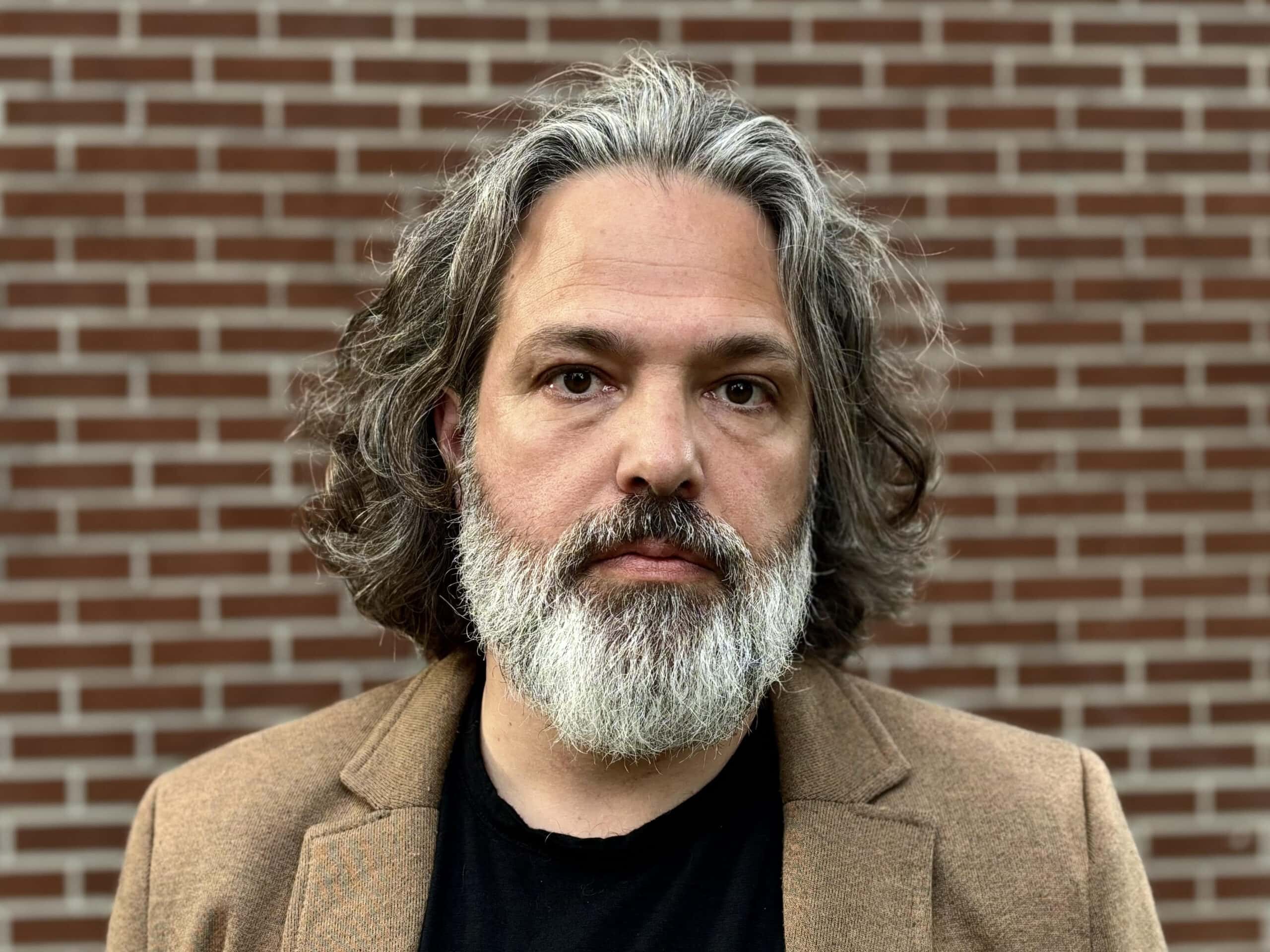 Thoughtful man with grey hair and beard, dressed in a brown blazer, standing in front of a brick wall, representing professionalism and style.