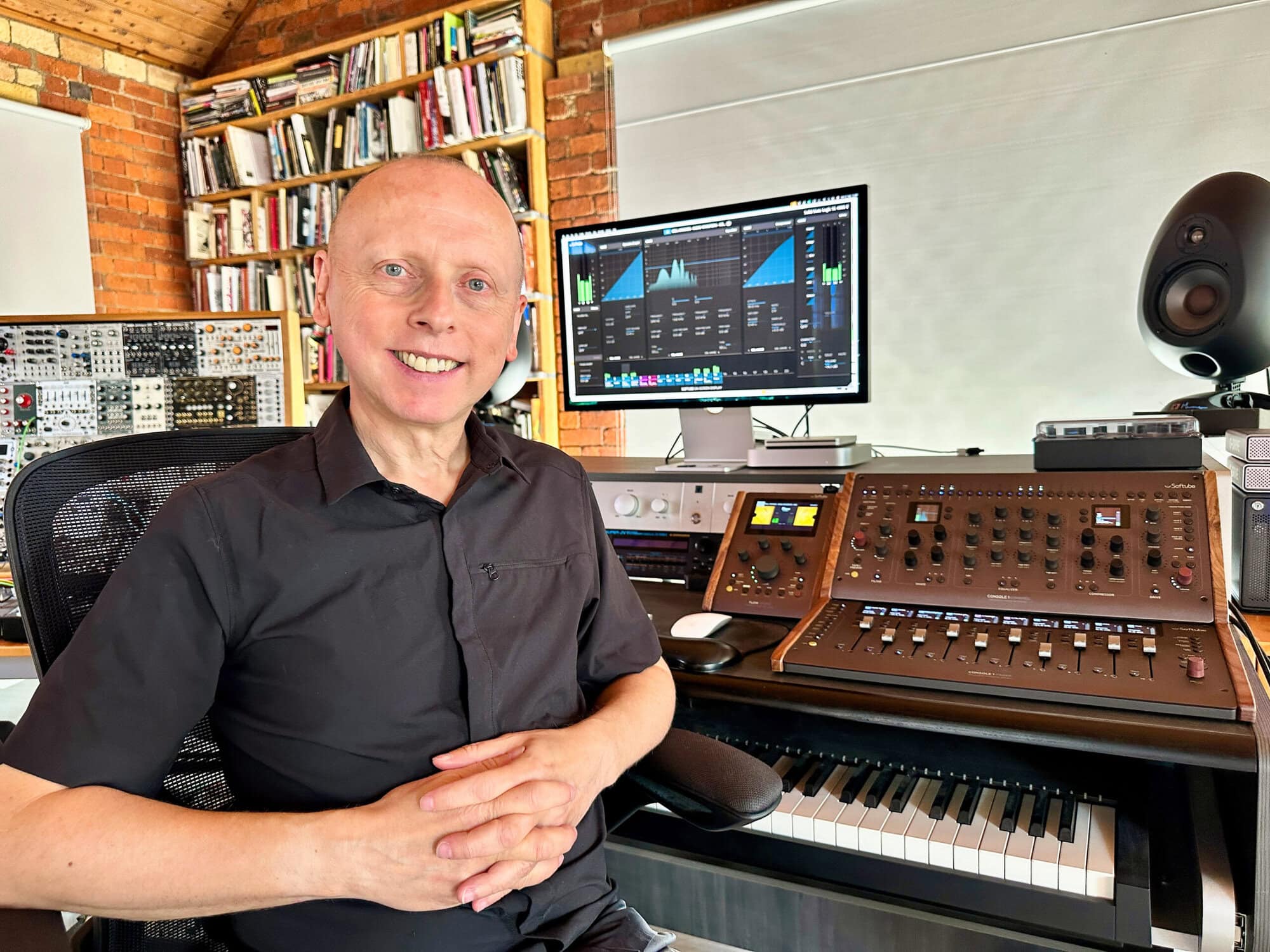A man in a black shirt smiling in a music studio with electronic equipment and a computer screen displaying audio editing software.