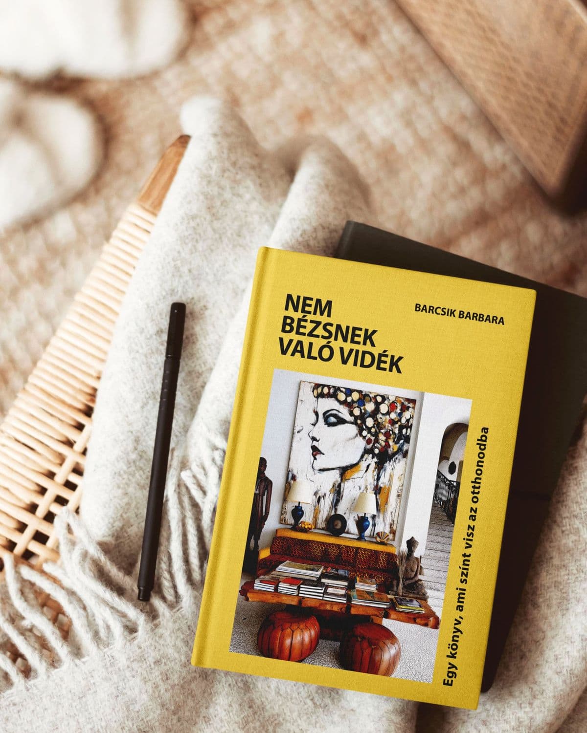 A Nem bézsnek való vidék könyv krémszínű gyapjúplédon, rattan szék mellett, egy fekete tollal