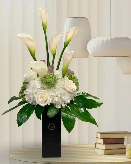Elegant white floral arrangement with calla lilies, roses, and hydrangeas in a sleek black vase for sophisticated decor or special occasions.