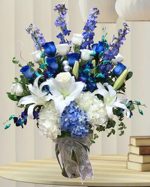 Blue and white floral arrangement featuring roses, lilies, hydrangeas, and delphiniums in a clear glass vase with elegant ribbon, perfect for special occasions.
