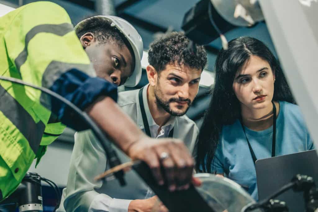 Three diverse apprentices in high-visibility safety gear and business attire collaborating around industrial equipment, demonstrating hands-on learning and teamwork in business administration apprenticeship training environment.