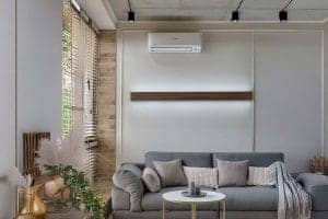 An image of an air conditioning unit on the wall of a modern looking living room. The air conditioning unit is situated above a grey sofa
