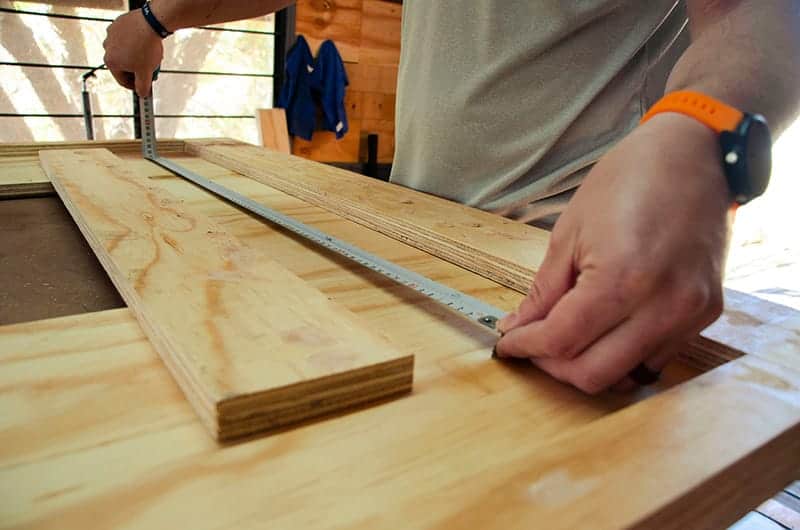 A close up of a carpenters hands measuring lengths of plywood with a tape measure