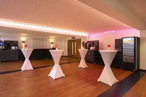 CongressCentrum Foyer – Diagonale Perspektive. Vier Stehtische mit Hussen, Catering-Bereiche links und rechts mit Kaffeemaschine und Getränkekühlschrank, geschlossene Doppeltür zum Saal, farbige Lichtstimmung, Holzboden
