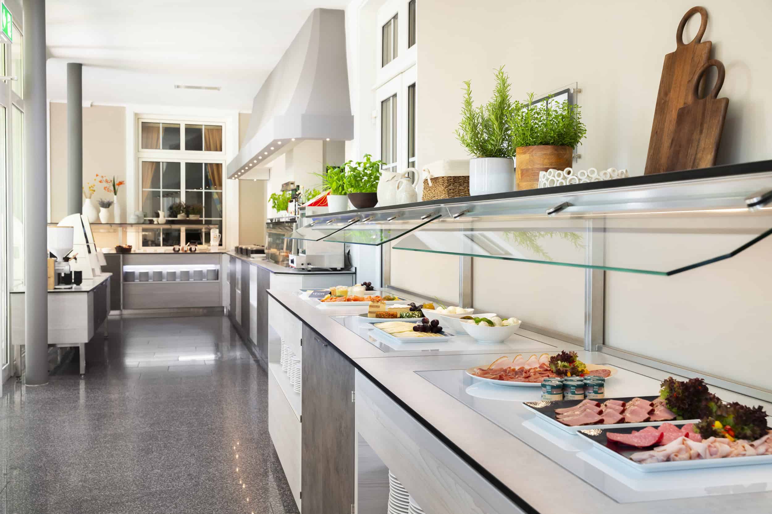 Moderne Frühstücksbuffet-Gesamtansicht: Aufschnitt- und Käsestationen, Nussmus-Maschine, helle Atmosphäre