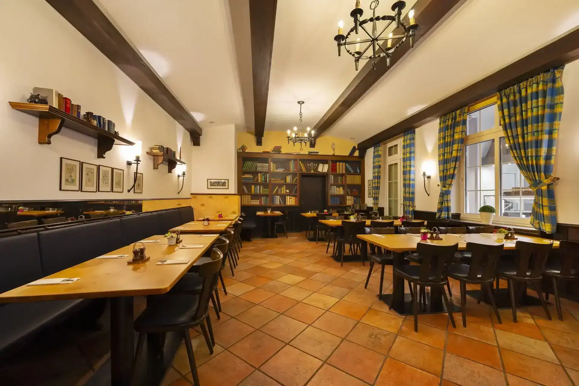 Kaminzimmer – diagonale Ansicht. Rustikale Restaurant-Bestuhlung mit Holztischen, Bücherregal-Tapete als Wandgestaltung hinten, sichtbare Holzbalken an der Decke, schmiedeeiserne Kronleuchter, Fenster mit karierten Vorhängen, terrakottafarbenem Fliesenboden
