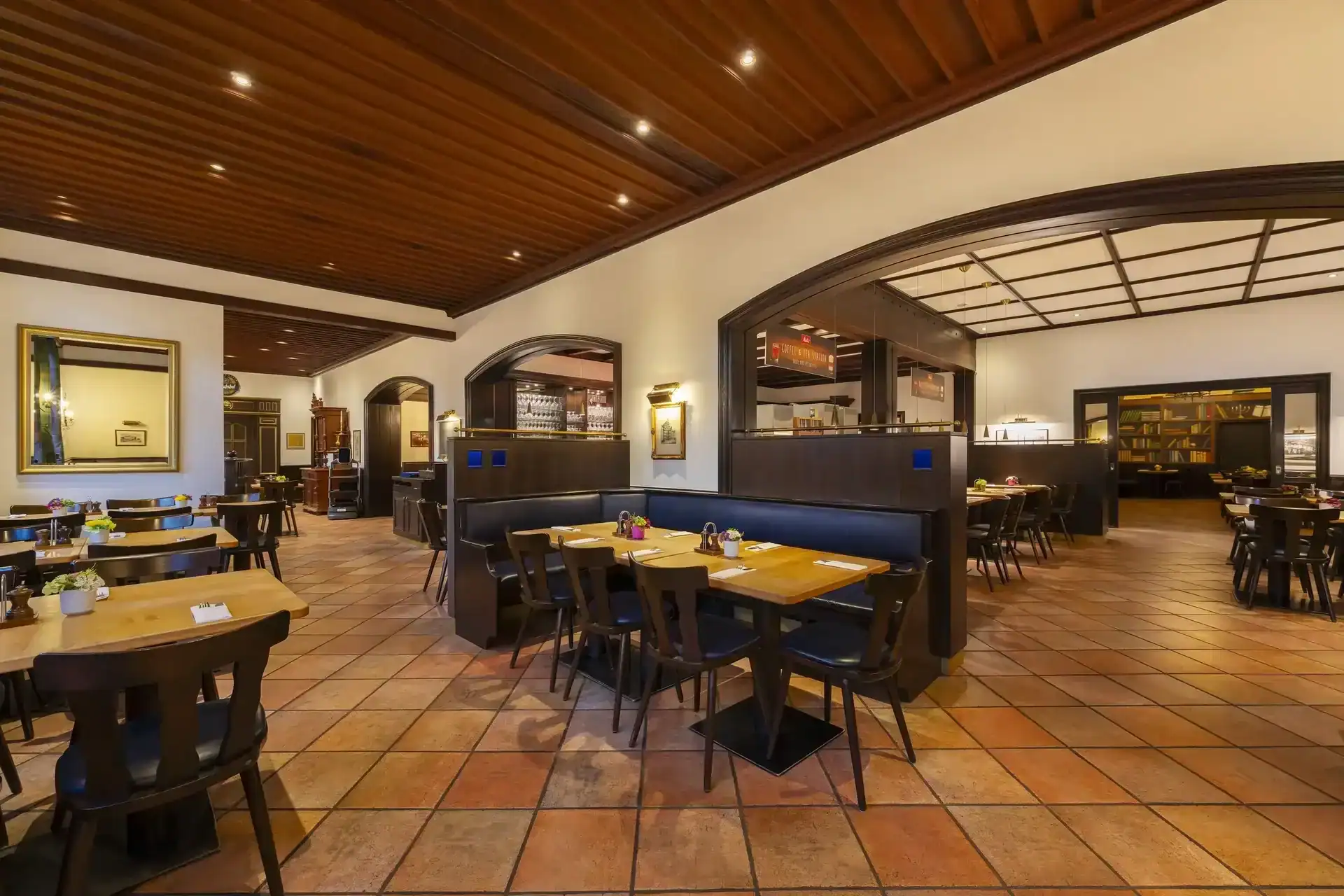 Restaurant – zentrale Ansicht. Rustikale Restaurant-Bestuhlung, Tische für 2-6 Personen, Sitzbänke als Raumteiler, Holzlamellendecke mit integrierten Spots, große Rundbogendurchgänge zu den verschiedenen Restaurantbereichen, terrakottafarbener Fliesenboden
