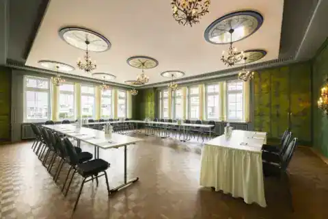 Spiegelzimmer – diagonale Ansicht. U-förmige Bestuhlung (bis 28 Personen), neun ornamentale Deckenrosetten mit floralen Kronleuchtern, historische grüne Wandgestaltung, Fensterfront an zwei aneinandergrenzenden Wänden, Parkettboden