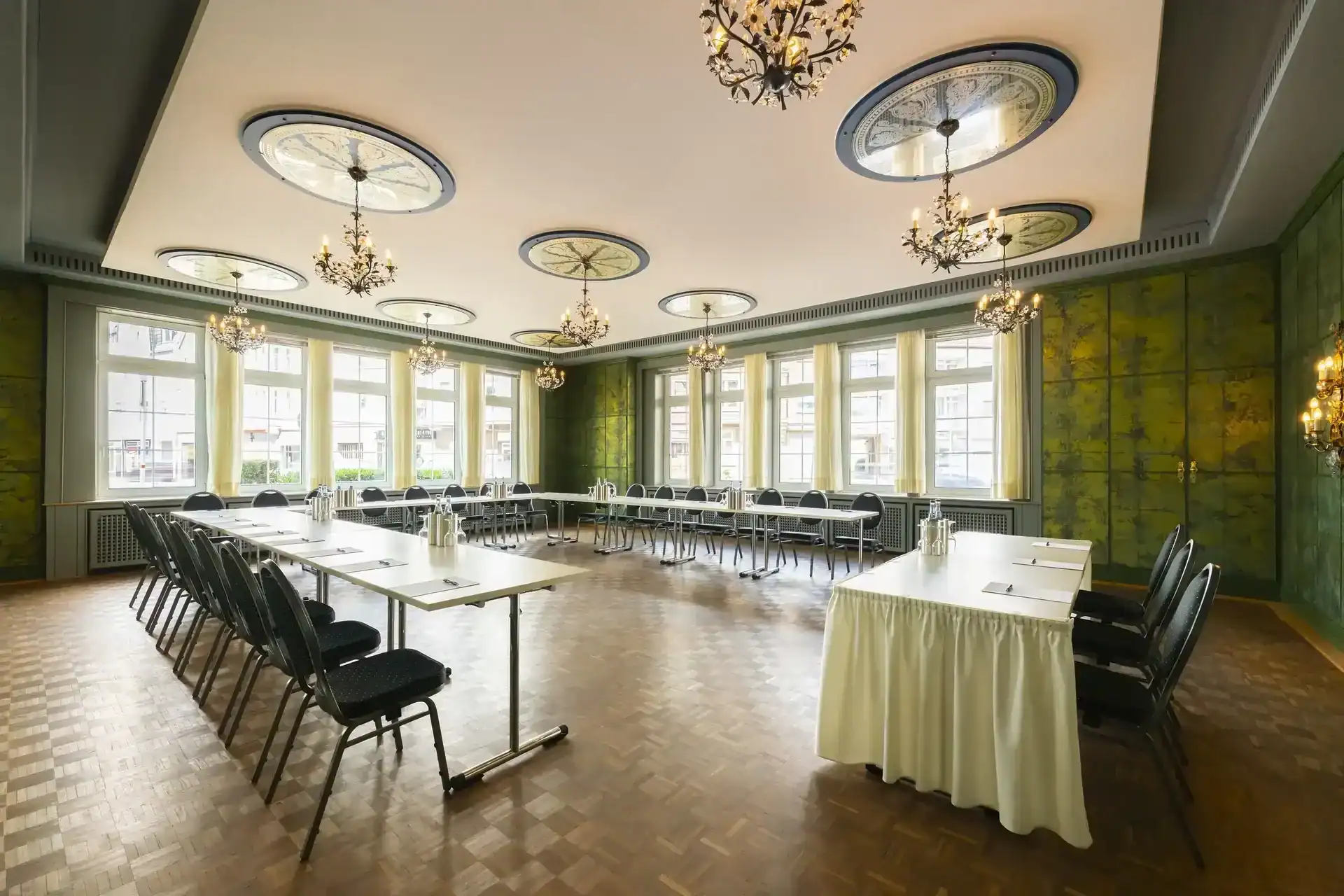 Spiegelzimmer – diagonale Ansicht. U-förmige Bestuhlung (bis 28 Personen), neun ornamentale Deckenrosetten mit floralen Kronleuchtern, historische grüne Wandgestaltung, Fensterfront an zwei aneinandergrenzenden Wänden, Parkettboden