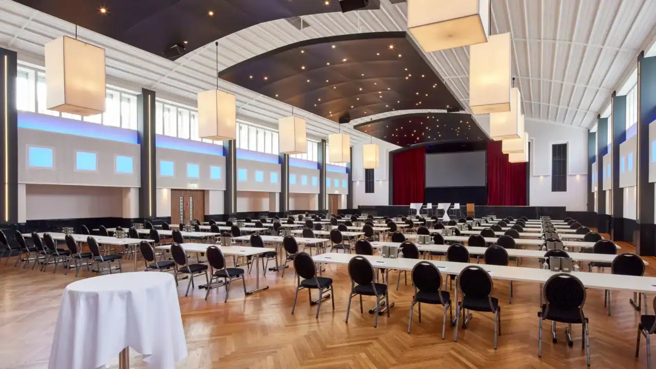 Großer Saal – zentrale Ansicht. Parlamentarische Bestuhlung, Bühne mit Leinwand und roten Vorhängen, gewölbte Deckenelemente mit Sternenhimmel-Beleuchtung, rechteckige Pendelleuchten, Fischgrätparkett