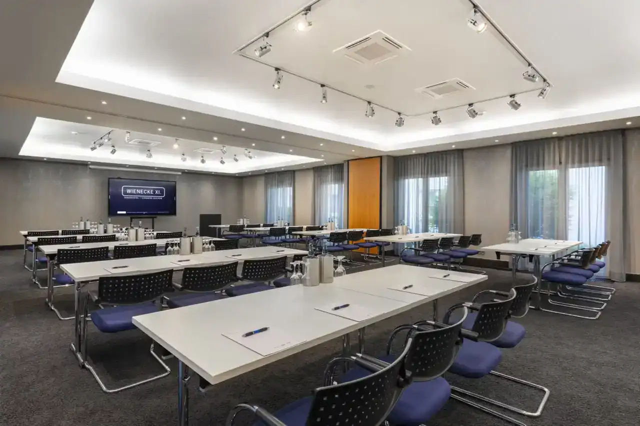 Tagungsraum Hotel – diagonale Ansicht nach vorne. Parlamentarische Bestuhlung für 40 Personen, mit blauen Freischwingern, LED-Display an Stirnwand, zwei eingelassene Deckenbereiche mit indirekter Beleuchtung und verstellbaren Strahlern, bodentiefe Fenster rechts mit Vorhängen, Teppichboden