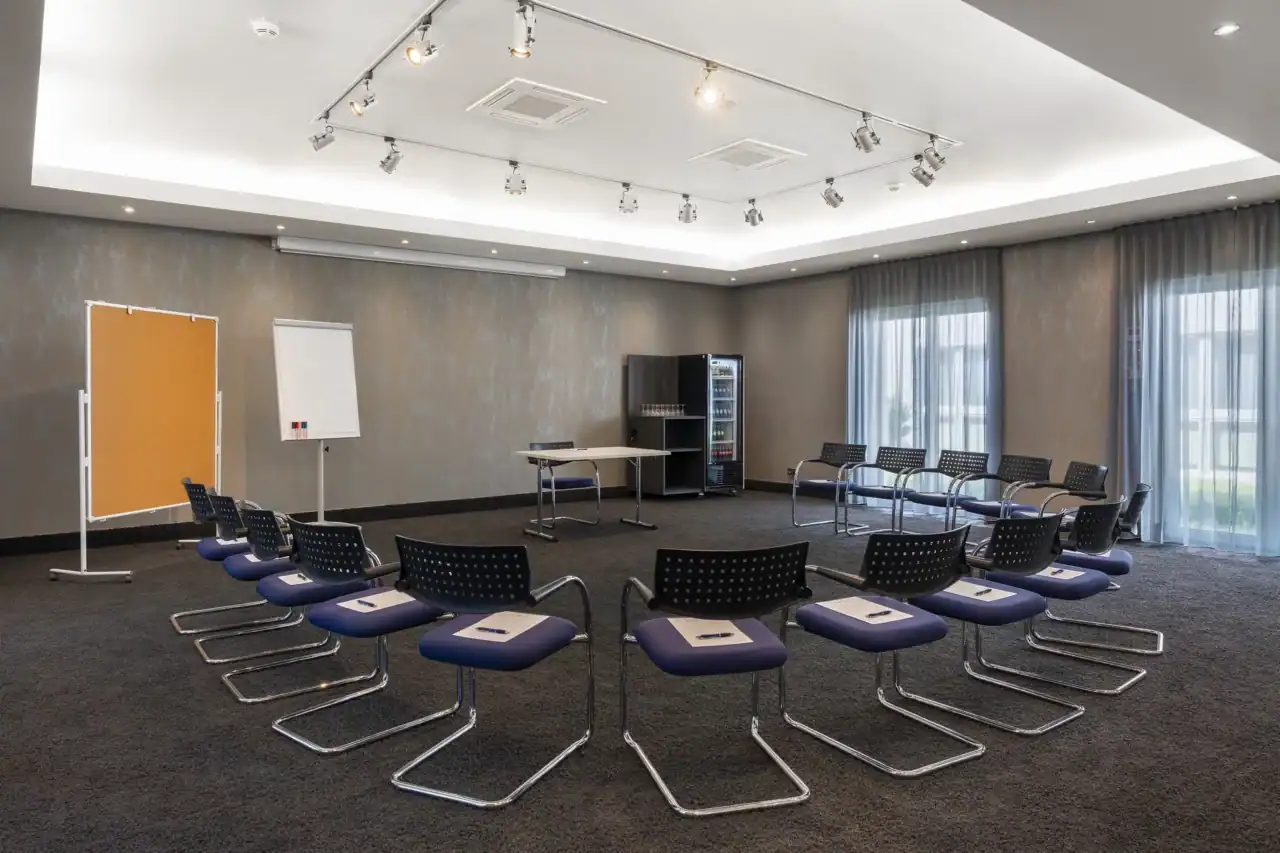 Tagungsraum II – Diagonale Perspektive: Im Zentrum Stuhlkreis-Bestuhlung, mit Freischwingern und einem Tisch. Self-Service Getränkekühlschrank in der gegenüberliegenden Raumecke. Flipchart und Pinnwand links, zwei bodentiefe Fenster rechts, verschiedene Deckenstrahler