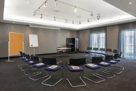 Tagungsraum II – Diagonale Perspektive: Im Zentrum Stuhlkreis-Bestuhlung, mit Freischwingern und einem Tisch. Self-Service Getränkekühlschrank in der gegenüberliegenden Raumecke. Flipchart und Pinnwand links, zwei bodentiefe Fenster rechts, verschiedene Deckenstrahler