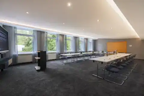 Tagungsraum VII – Blick Rückwand zu Eingangstür aus Holz. U-Form Bestuhlung mit Konferenztischen und 24 Freischwingern, Display links, Rednerpult mit Stauraum, Fensterfront, Teppichboden