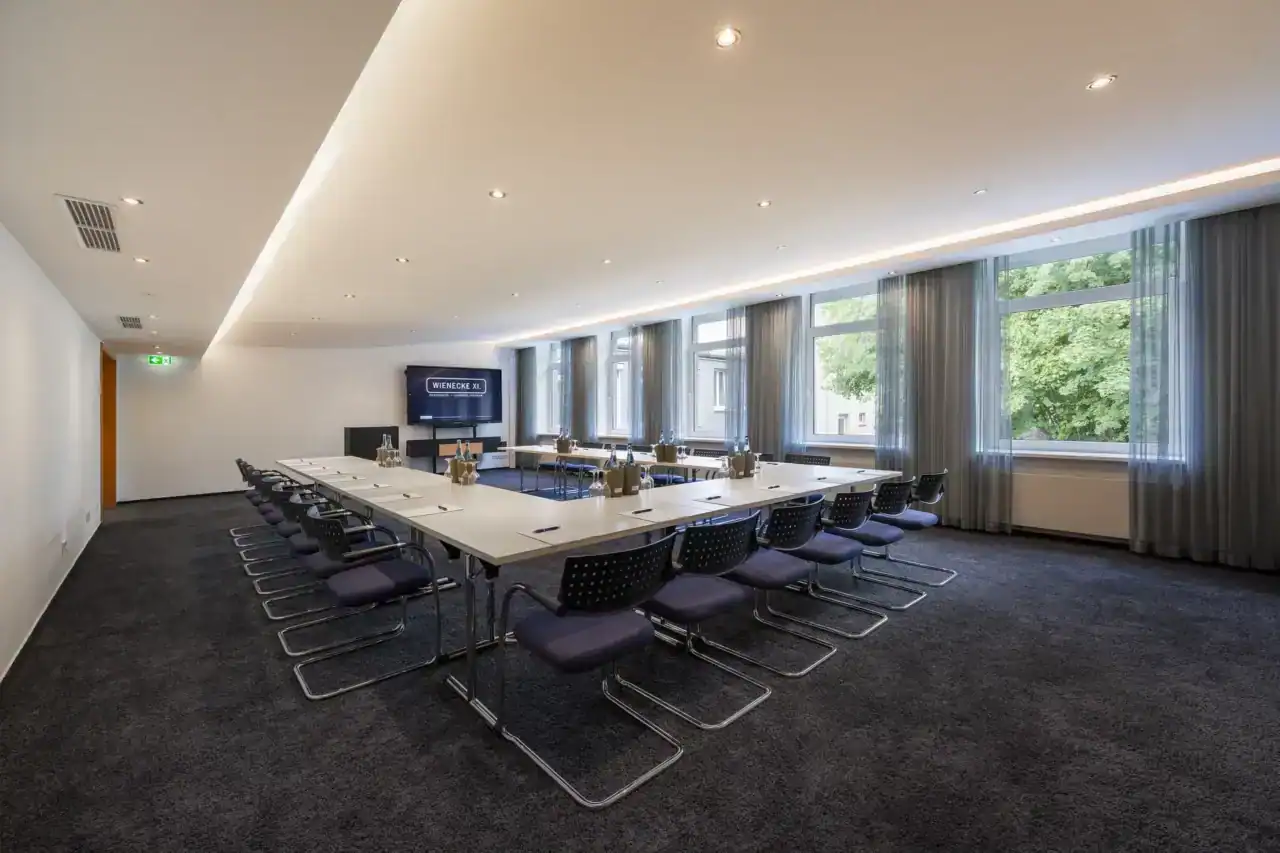 Tagungsraum VII – Diagonale Perspektive. U-Form Bestuhlung mit Konferenztischen und 24 Freischwingern, Rednerpult, mobiles Display, Fensterfront (5 Fenster), indirekte Deckenbeleuchtung, Teppichboden