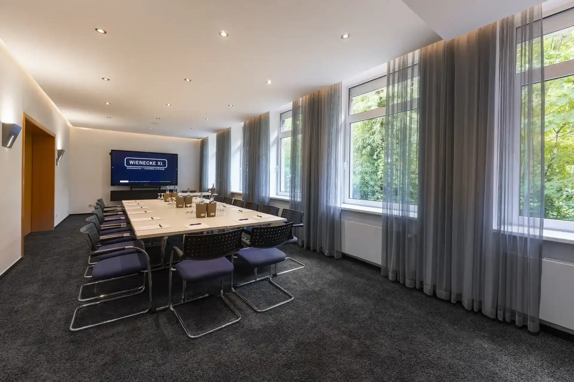 Tagungsraum VIII – Diagonale Perspektive. Konferenztisch mit 14 Freischwingern, Display an der Wand, Fensterfront mit Blick ins Grüne, Verbindungstür links, indirekte Deckenbeleuchtung, Teppichboden