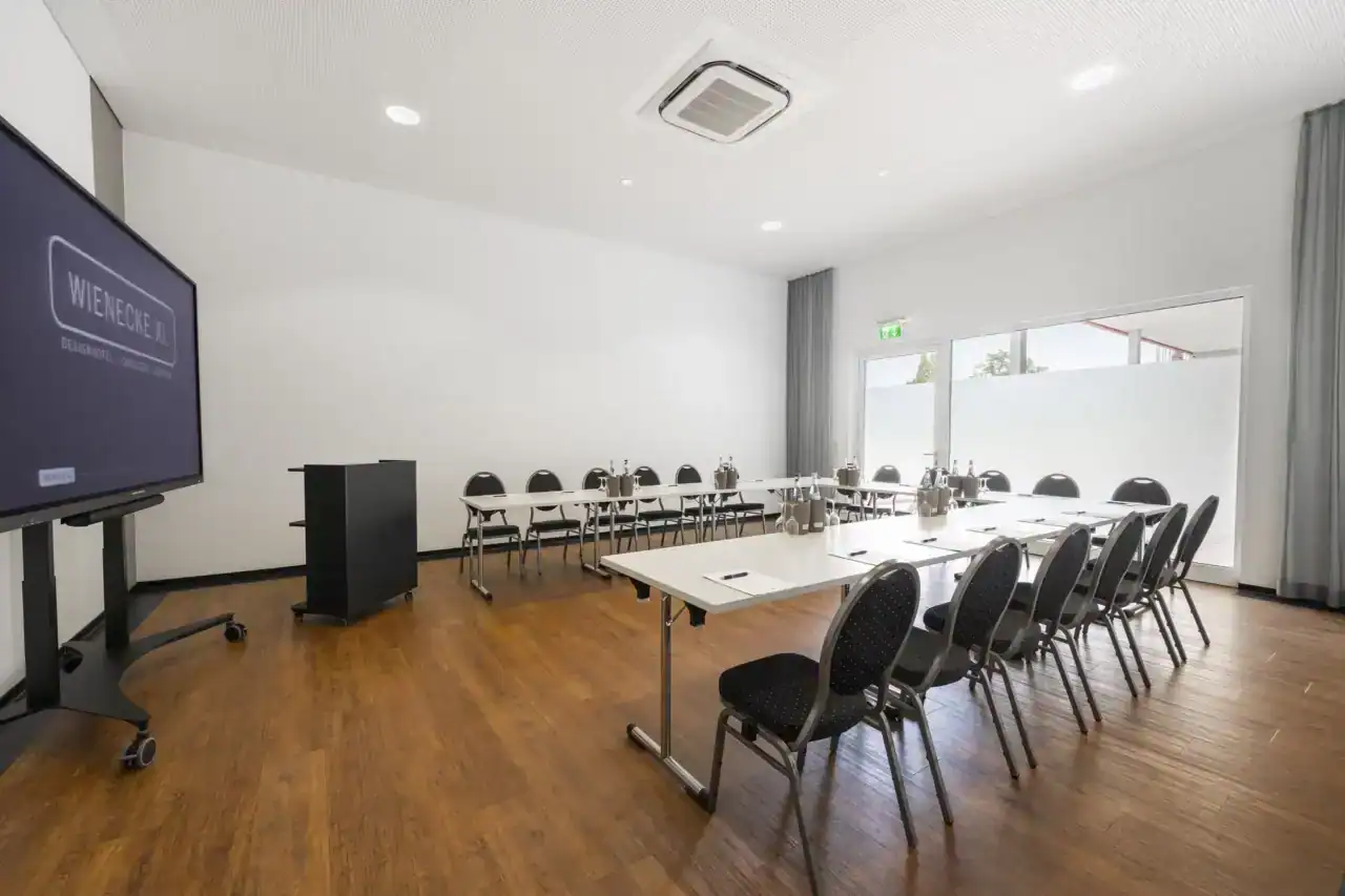 Tagungsraum XX – Diagonale Rückansicht. U-Form Bestuhlung mit 18 Polsterstühlen, mobiles Display links, Rednerpult, Holzboden, bodentiefe Fenster mit Sichtschutzfolie