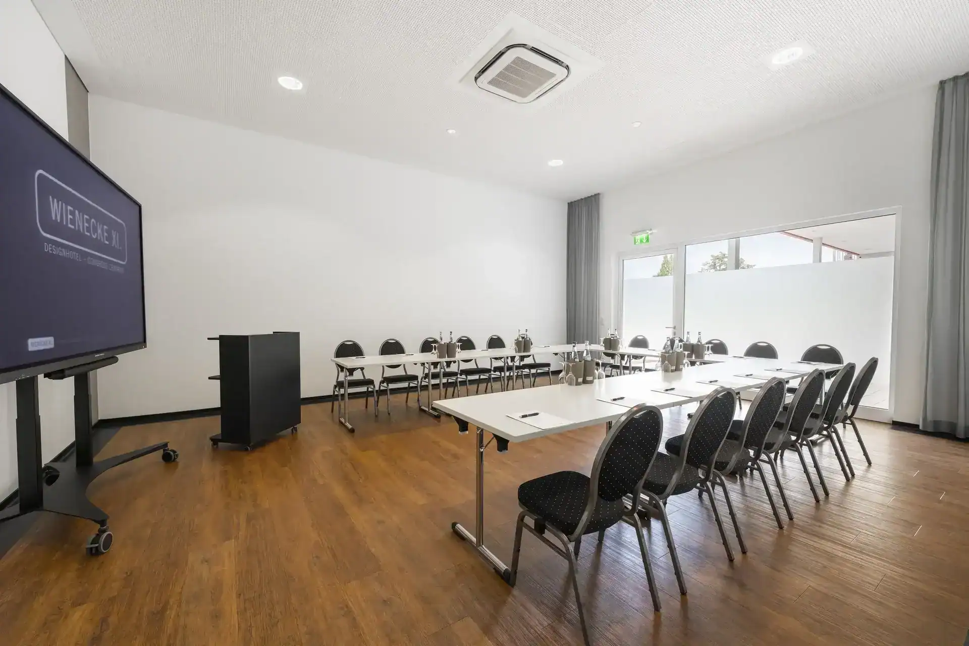Tagungsraum XX – Diagonale Rückansicht. U-Form Bestuhlung mit 18 Polsterstühlen, mobiles Display links, Rednerpult, Holzboden, bodentiefe Fenster mit Sichtschutzfolie