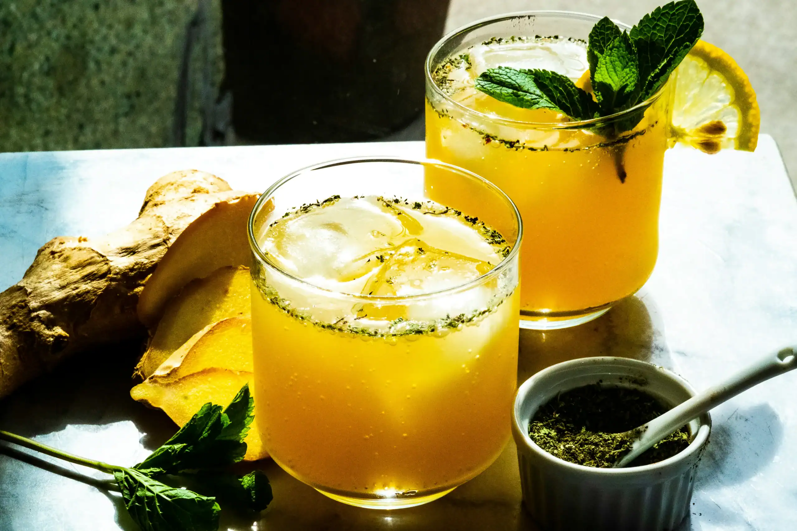 Two glasses filled with digestive bitters for digestion sit on a table amidst ginger root and herbs
