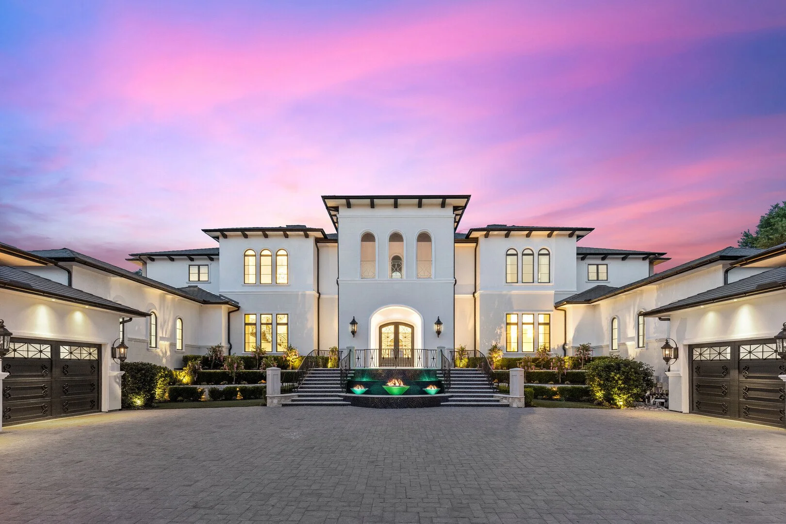 Luxurious Mediterranean-style mansion with arched entryway, balconies, and landscaped exterior illuminated at sunset.