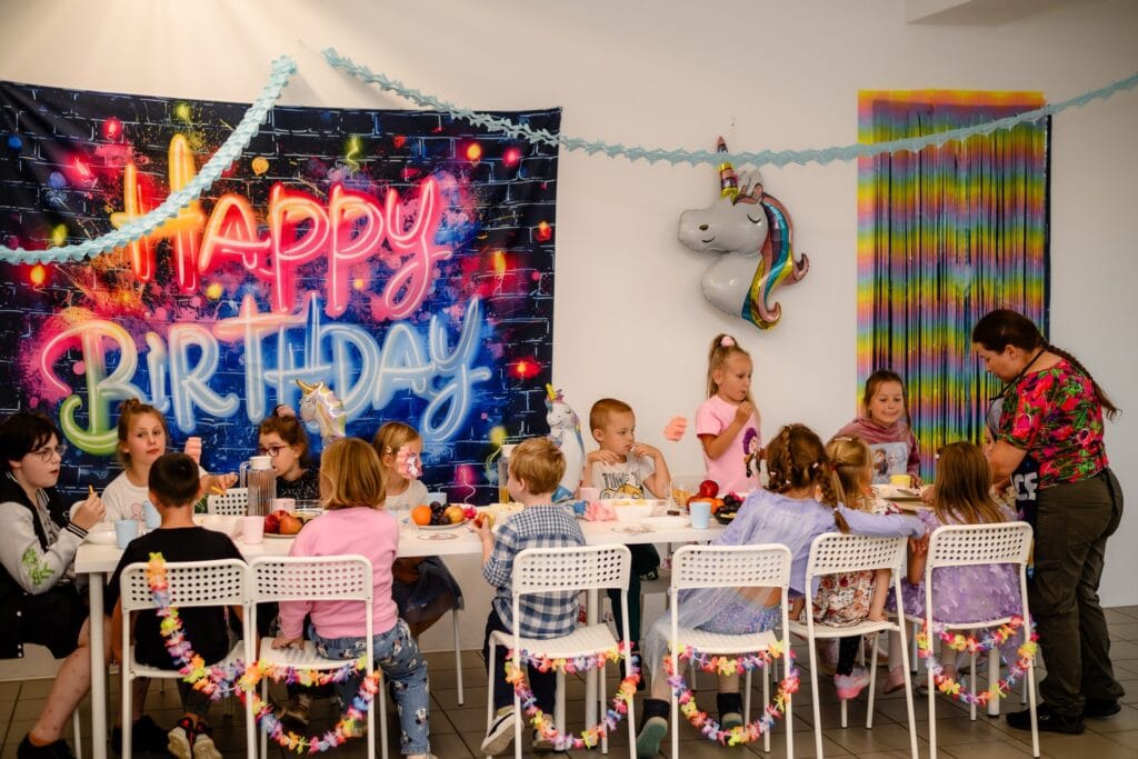 Kolorowa impreza urodzinowa dla dzieci, dekoracje w stylu Disney i balony na tle neonowego napisu Happy Birthday, dzieci bawią się przy stole, w centrum atmosfera radości, kolorowe ozdoby i motyw unicorn.