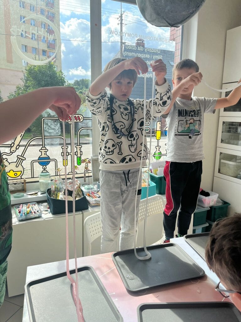 RYS. Dzieci uczestniczące w warsztatach naukowych, eksperymentując z chemikaliami, z laboratoryjnym wyposażeniem i kolorową dekoracją, promujące edukację i naukowe pasje najmłodszych.