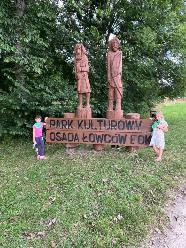 Pomniki drzew liściastych w parku kulturowym Osada Łowców Fok wśród zieleni i drzew, dzieci stojące obok, duży drewniany napis informujący o miejscu, naturalne otoczenie, miejsce edukacji i rekreacji w kontakcie z naturą.