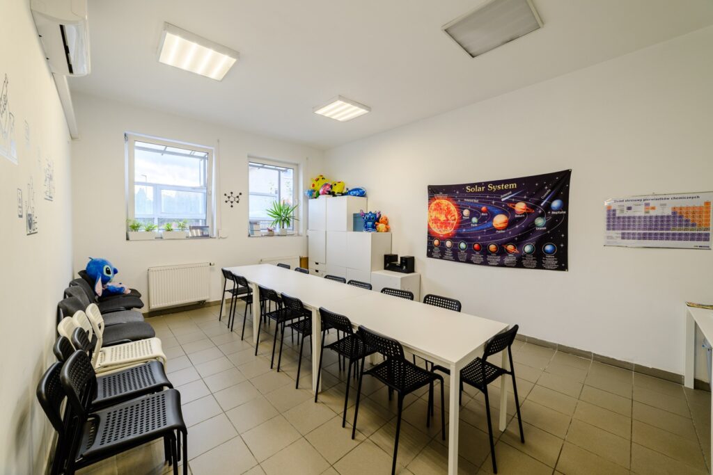 Kolorowa sala szkoleniowa z dużym stołem, krzesłami, plakatem systemu słonecznego i tablicami edukacyjnymi, wypełniona zabawkami, roślinami i pomieszczeniem edukacyjnym dla dzieci i studentów.
