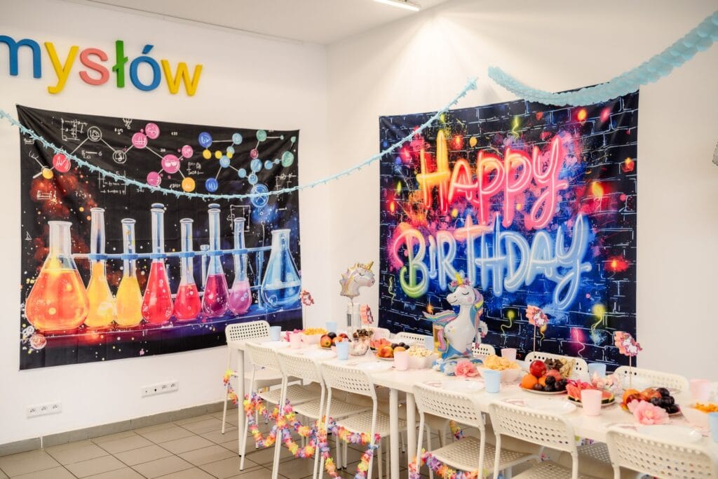 Kolorowy baner z chemicznymi probówkami i neonowe napisy "Happy Birthday" na tle ściany z elementami nauki, idealne do dekoracji urodzinowej dla dzieci z motywem nauki i magii.