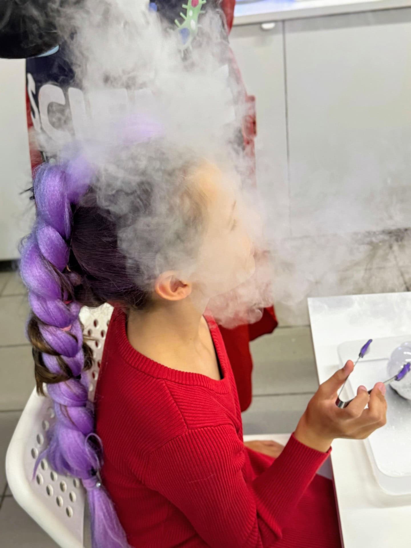 Foka z kolorowym warkoczem podczas eksperymentu naukowego z dymem suchym azotem w laboratorium. W tle widoczne elementy związane z technologią i nauką, idealne do promowania innowacyjnych rozwiązań edukacyjnych.
