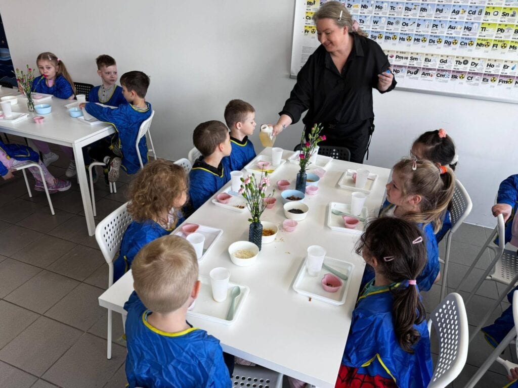 Nauczycielka przeprowadzająca zajęcia edukacyjne z grupą dzieci w klasie szkolnej, podczas warsztatów kulinarnych, z tablicą z chemicznymi symbolami pierwiastków na tle, w przedszkolu lub szkole.
