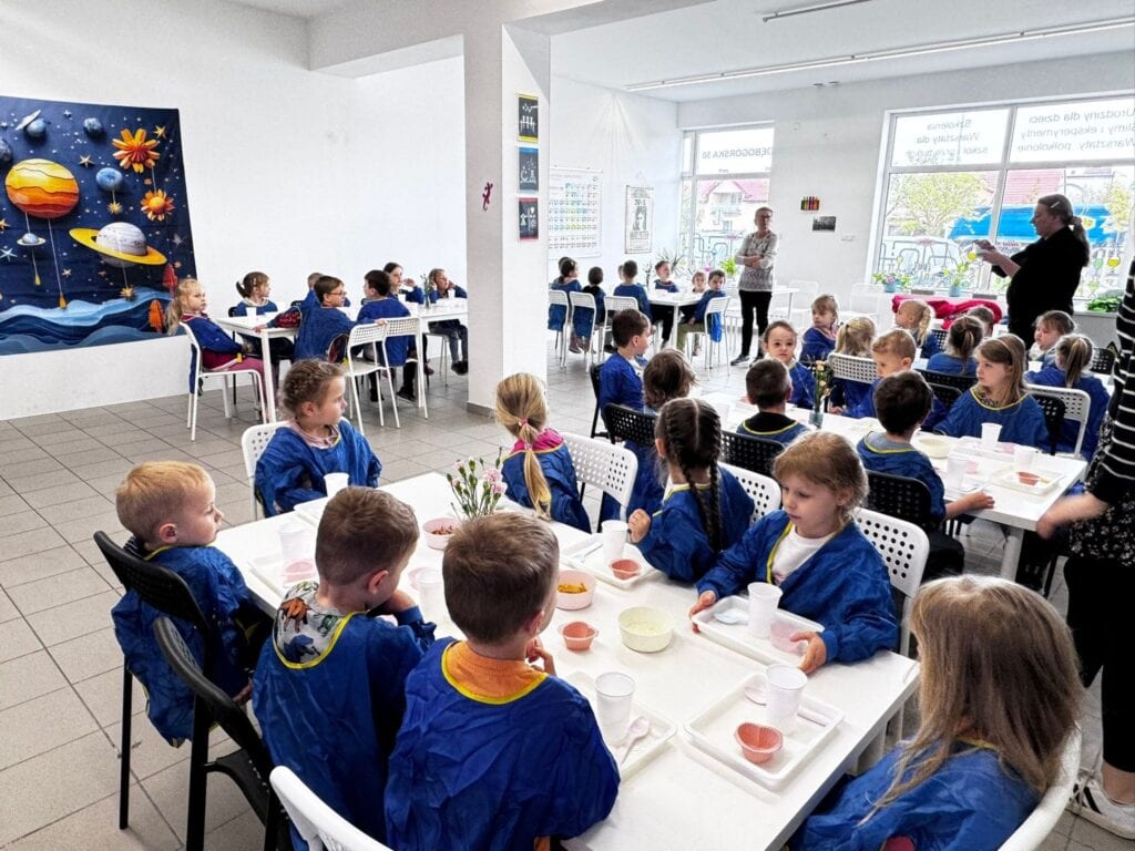 Edukacyjna przestrzeń dla dzieci podczas zajęć w nowoczesnej szkole, z uczniami w niebieskich fartuchach, prowadzone przez nauczycielkę w jasnym swetrze, w jasnym wnętrzu z dużymi oknami i dekoracjami edukacyjnymi.