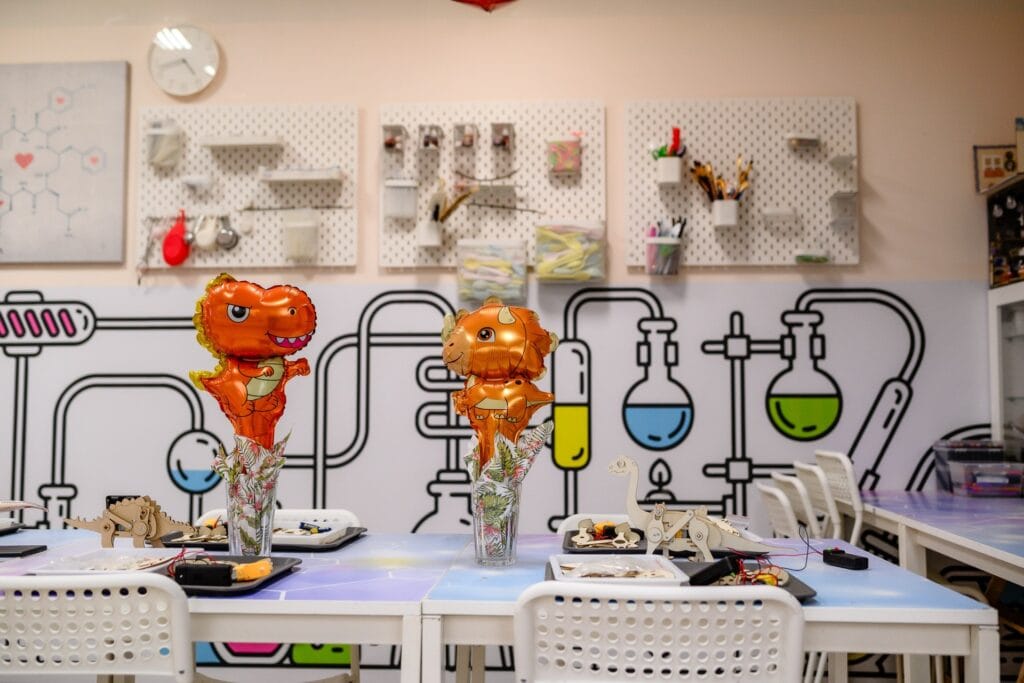 Kolorowe balony w kształcie dinozaurów na tle naukowo-inżynieryjnego wnętrza w pracowni Wittlab, gdzie tworzy się innowacyjne projekty edukacyjne i naukowe dla dzieci i młodzieży.