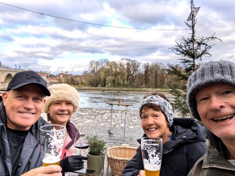 Shared memories on a Tauck Small Ship Cruise. Cruise participants enjoying drinks by the riverside in a scenic European town, showcasing travel experiences with friends, picturesque waterfront, and casual outdoor dining in a charming setting.