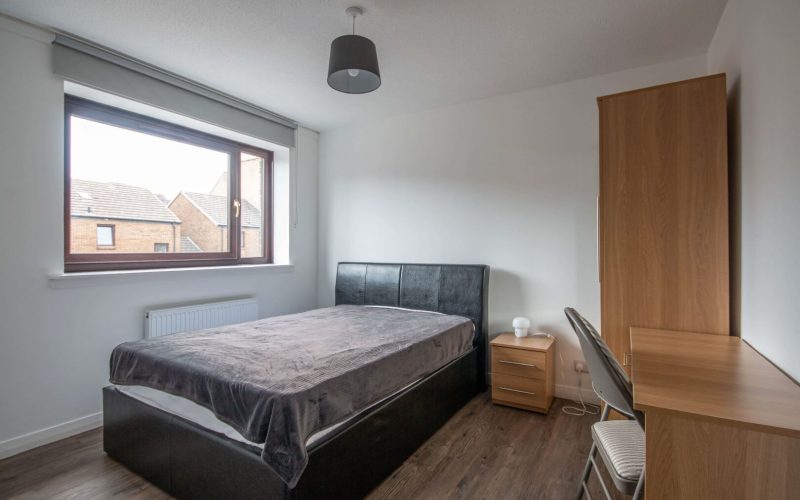 Comfortable guest bedroom featuring a wooden wardrobe, desk, and large window showcasing neighbourhood rooftops.