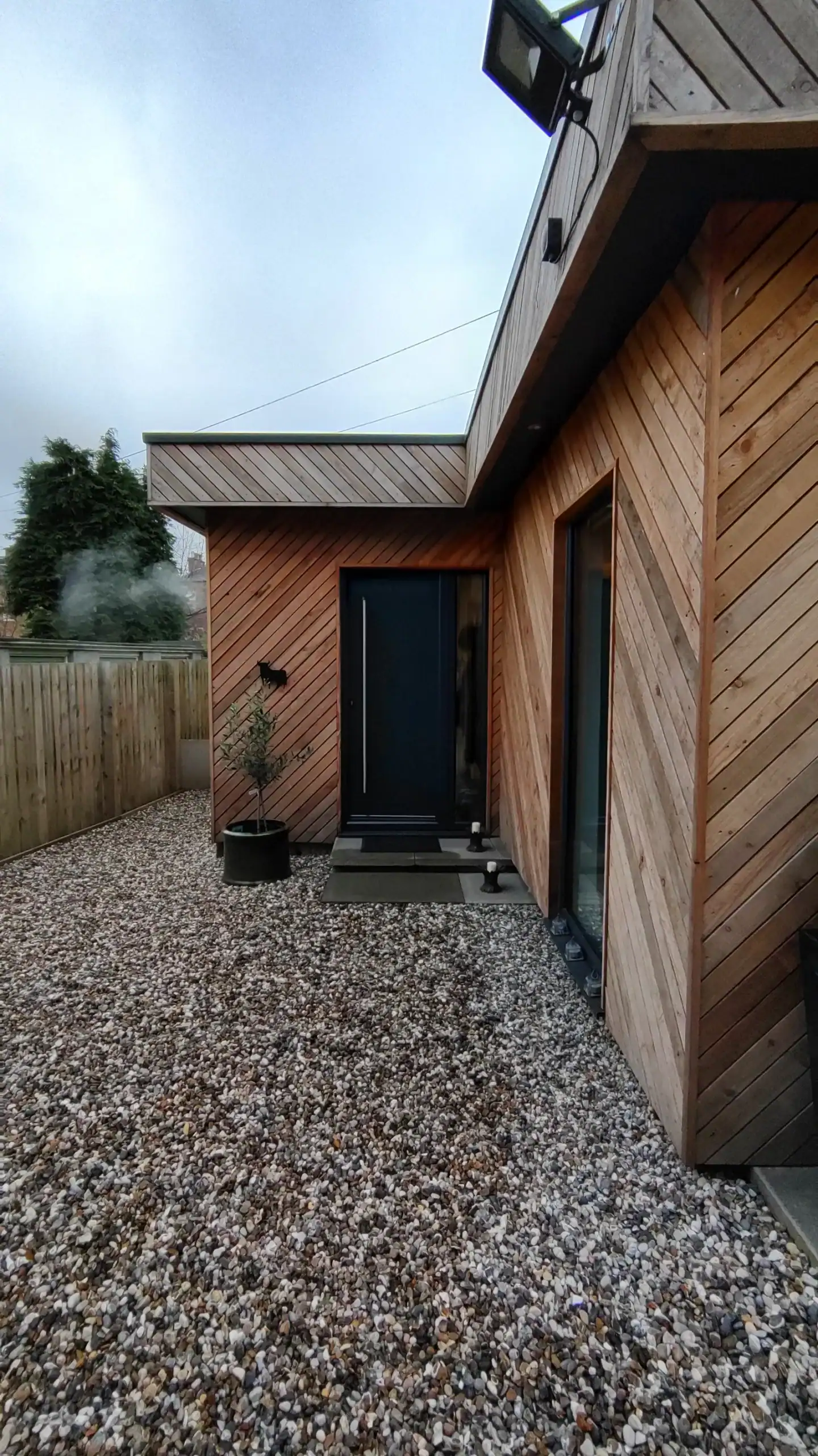 Alt: Contemporary wooden house exterior with gravel pathway and black door, eco-friendly architecture.