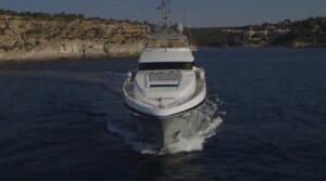 Un yacht de luxe blanc navigue sur une eau bleu foncé, créant un sillage derrière lui. Des falaises rocheuses à la végétation clairsemée et quelques bâtiments sont visibles le long du littoral – comme si l'on cherchait des trésors cachés sous un ciel dégagé.