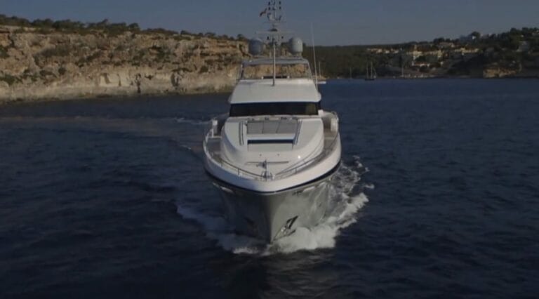 Un yacht de luxe blanc navigue sur une eau bleu foncé, créant un sillage derrière lui. Des falaises rocheuses à la végétation clairsemée et quelques bâtiments sont visibles le long du littoral – comme si l'on cherchait des trésors cachés sous un ciel dégagé.