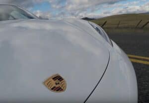Close-up of the front left of a silver Porsche, with the Porsche emblem on the bonnet. This stunning car drives down a deserted rural road in Quebec, bordered by grassy hills and a fence, under a cloudy blue sky. 