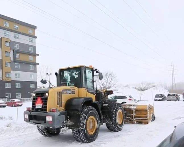 Une chargeuse frontale jaune équipée d'un chasse-neige déneige un parking entouré de congères. Des voitures sont garées à proximité et un immeuble d'appartements se dresse sur la gauche. Des lignes électriques sont visibles au-dessus, signe que les défis hivernaux seront relevés de front.