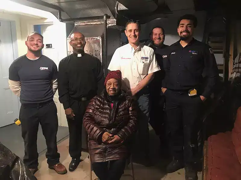 A seated woman in a red coat is surrounded by five standing men in work uniforms and a clergy outfit, posing for a photo indoors.