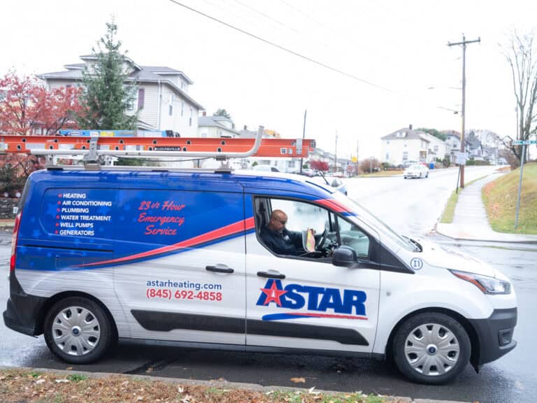 A blue and white Astar service van is parked on a suburban street with a person seated inside; a ladder is secured on the van's roof.